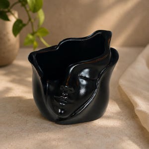 Könnte beinhalten: Ein schwarzer Keramikübertopf mit einem skulptierten menschlichen Gesicht mit geschlossenen Augen und einem ruhigen Ausdruck. Das Gefäß hat einen unregelmäßigen, gewellten Rand und eine glatte, glänzende Oberfläche für kleine Pflanzen.