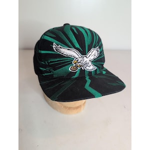 May include: A black baseball cap featuring a vibrant green sunburst pattern radiating from the center. An embroidered white eagle with outstretched wings is centered on the front panel, clutching a small brown football in its talons.