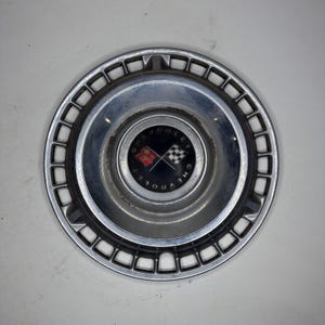 May include: A circular chrome Chevrolet hubcap featuring a central black emblem with the text CHEVROLET, a red bowtie logo, and a checkered flag. The outer rim has a series of rectangular cutouts arranged in a ring around the polished metal surface.