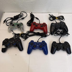 Puede incluir: Una colección de seis mandos de juego con cable en varios colores, incluyendo transparente, rojo, negro y azul. Cada mando incluye una cruceta, dos joysticks analógicos y varios botones de acción, todos conectados mediante cables de color negro.
