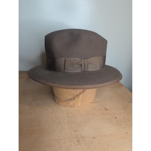 May include: A dark brown fedora hat featuring a matching dark brown grosgrain ribbon band tied in a bow at the side. The hat has a structured crown with a centre crease and a wide, flat brim, showcasing a smooth, uniform texture throughout.