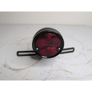May include: A vintage-style black tail light with two red lenses. The light has a circular shape with a metal bracket for mounting. The lenses have a textured pattern. The tail light is designed for a vehicle.