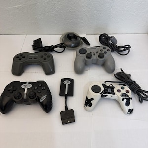 May include: A collection of four video game controllers and one adapter. The set includes a dark gray controller, a light gray controller with a cord, a black controller, a white controller with black splatters, and a black rectangular adapter.