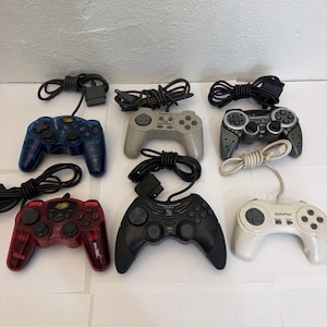 May include: A collection of six wired video game controllers featuring various designs, including translucent blue and red models, a grey controller, a black controller labelled Storm Chaser, a dark grey model, and a white controller labelled GamePad.