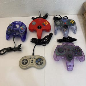 Puede incluir: Una colección de cinco mandos de videojuegos que incluye un mando morado translúcido, un mando rojo sólido, un mando gris translúcido, un mando gris con cuatro botones y un mando morado translúcido grande, todos con cables negros.