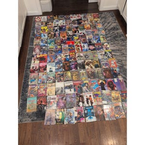 May include: A large collection of comic books, featuring various titles and colorful covers, is spread out on a rug. The comics are arranged in a grid-like pattern, showcasing a variety of artwork and logos. The collection includes a diverse range of genres and publishers.