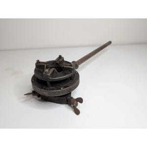 May include: An antique, dark metal boat motor with a propeller and a long, cylindrical handle. The motor has a circular base with a series of bolts and a star-shaped top. The propeller has three blades. The object is set against a white background.