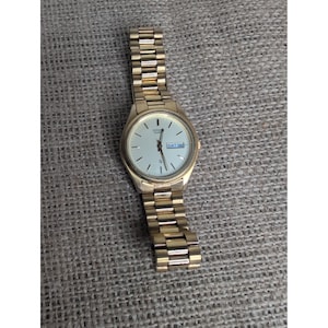 Pode incluir: Um relógio de pulso Citizen Quartz em tom dourado com mostrador redondo branco, marcadores de hora pretos finos, ponteiros dourados e uma janela de dia e data na posição das três horas exibindo o texto SAT 16. Possui uma pulseira de elos metálicos.