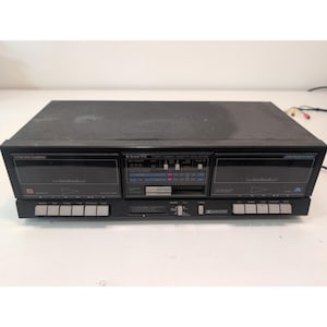 Puede incluir: Una pletina de casete estéreo doble Sanyo de color negro con dos compartimentos para cintas etiquetados como A y B, un panel de control central con contador de cinta, medidores de nivel y varios botones plateados para funciones de transporte.