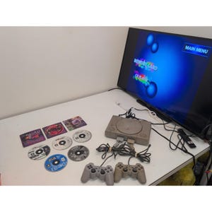May include: A gray video game console with two matching controllers, several game discs, and a television screen displaying a blue main menu interface with the text MAIN MENU, MEMORY CARD, and CD PLAYER. The console is connected to the television.