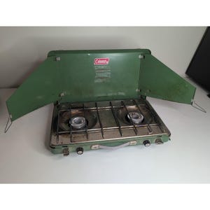 Puede incluir: Una estufa de camping Coleman verde vintage con dos quemadores. La estufa tiene un marco de metal y un protector contra el viento verde que se pliega. El logotipo de Coleman es visible en la parte delantera. La estufa está diseñada para cocinar al aire libre.