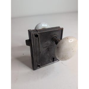 Op de afbeelding: Een metalen insteekslotmechanisme met een rechthoekige donkergrijze plaat met een sleutelgat en een krukstift. Twee ronde, gebroken witte porseleinen deurknoppen zijn aan de stift bevestigd en vertonen duidelijke tekenen van ouderdom en slijtage.