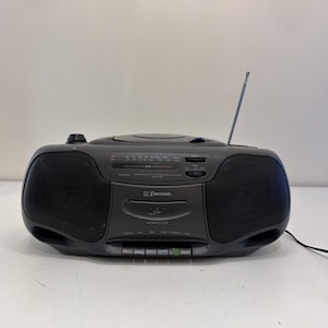 Op de afbeelding: Een antracietgrijze draagbare Emerson cassettespeler en radio met een cd-compartiment aan de bovenzijde, dubbele luidsprekers, een uitschuifbare metalen antenne en een bedieningspaneel aan de voorzijde voor afspelen en radio-afstemming.