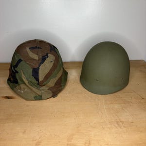 Puede incluir: Dos cascos de estilo militar se muestran uno al lado del otro. Un casco presenta una funda de tela con patrón de camuflaje boscoso en tonos marrón, verde y negro. El segundo casco es de color verde oliva mate sólido con una forma redondeada.