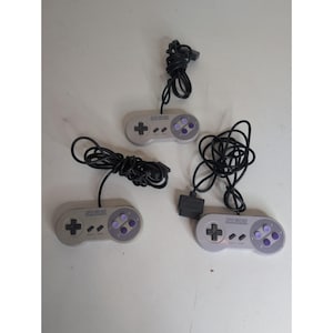 May include: Three grey Super Nintendo game controllers, each featuring a black directional pad, two small grey start and select buttons, and four purple action buttons on the right side. Each controller includes a long black wired connection cable.
