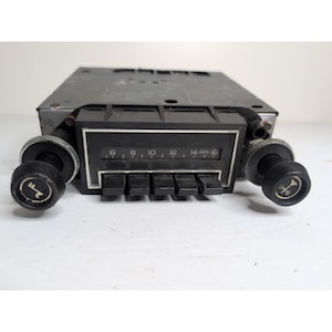 May include: A vintage car radio with a black faceplate and knobs. The radio features a tuning dial with numbers and a series of push buttons. The knobs have a symbol on them. The radio is set against a white background.