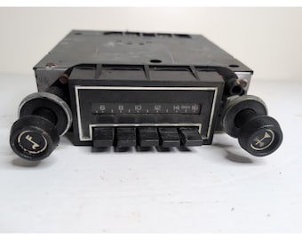 May include: A vintage car radio with a black faceplate and knobs. The radio features a tuning dial with numbers and a series of push buttons. The knobs have a symbol on them. The radio is set against a white background.