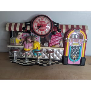 Könnte beinhalten: Eine Wanduhr mit einer Diner-Szene im Stil der 1950er Jahre, inklusive Coca-Cola-Logo, Jukebox und Theke mit Figuren. Das Zifferblatt ist rund mit roten Zahlen. Die Szene umfasst eine rot-weiß gestreifte Markise, eine Speisekarte und Barhocker.