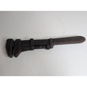 May include: A vintage adjustable wrench, primarily dark gray with a brown handle. The wrench features a movable jaw, a threaded adjustment mechanism, and a handle with two rivets and a diamond-shaped emblem.