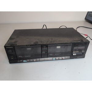 Puede incluir: Una pletina de casete estéreo doble Technics de color negro con dos compartimentos para cintas etiquetados como Deck A y Deck B, un botón de encendido, una toma de auriculares y una serie de botones de control en el borde inferior.