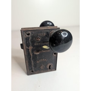 Puede incluir: Una cerradura de embutir antigua con una placa frontal rectangular de metal oscuro, orificios para tornillos y una cerradura. La placa tiene grabado el texto CORBIN y fechas de patente. Dos pomos negros, redondos y lisos, están acoplados.