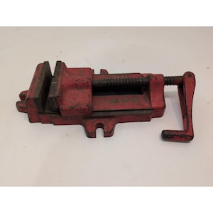 May include: A red metal bench vise featuring a heavy-duty cast iron body with visible wear and paint chips. The tool includes a threaded screw mechanism, a sliding jaw, and a manual crank handle for tightening and securing workpieces.