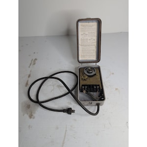 May include: A vintage industrial electrical timer housed in a rectangular metal case with a hinged lid. The interior features a circular dial, brass gears, and terminal connections. A black power cord with a two-prong plug is attached to the base.