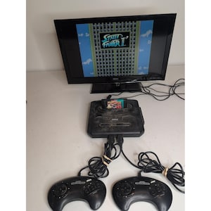 Pode incluir: Uma consola de videojogos Sega Genesis preta com dois comandos com fios e um cartucho de jogo Street Fighter II inserido. A consola está ligada a um televisor de ecrã plano que apresenta o ecrã de título de Street Fighter II.