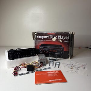 May include: A Sparkomatic SR600 compact disc player car stereo with its original box, wiring harness, metal mounting brackets, and an orange user manual. The black head unit features a digital display, volume knob, and various control buttons.