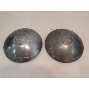 Puede incluir: Dos tapacubos circulares de metal plateado con el logotipo de Volkswagen en relieve en el centro. Estas cubiertas de rueda tienen una superficie lisa y abovedada con signos visibles de desgaste, incluyendo pequeños arañazos y oxidación.