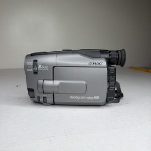 以下が含まれることがあります： シルバーのソニーHandycamビデオHi8ビデオカメラ。ビデオカメラは黒いレンズ、黒いストラップ、そして「30x SteadyShot」の文字があります。ビデオカメラには電源ボタンとフォーカスボタンがあります。