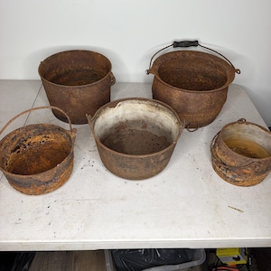 May include: A collection of five weathered cast iron pots and cauldrons featuring varying sizes and shapes. Each vessel displays a heavy rusted brown patina, with some including metal bail handles and one featuring a light-colored interior lining.