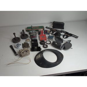 May include: A collection of vintage automotive parts, including gauges, mirrors, and engine components, arranged on a white surface. Various sizes and shapes are visible, with a mix of metal and other materials. The parts appear to be from different eras.
