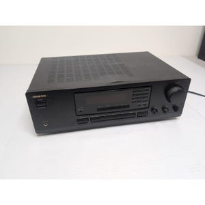 Pode incluir: Receptor estéreo Onkyo preto com visor digital, vários botões e um grande botão de volume. O receptor tem o nome da marca "ONKYO" impresso na frente.