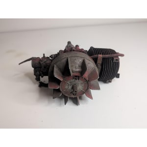 May include: A small, weathered internal combustion engine featuring a prominent circular cooling fan with multiple blades, a cylindrical finned engine block, and remnants of red paint on its metallic, industrial-style components.