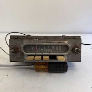 Puede incluir: Una radio de coche rectangular vintage con acabado gris metálico, que presenta un dial central marcado con los números 55, 6, 7, 9, 11, 13, 15, 16 y el texto All Transistor. Tiene cuatro botones rectangulares y dos perillas laterales sobre un destornillador.