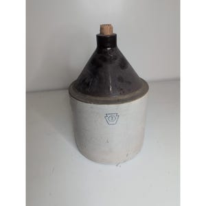 Puede incluir: Una jarra de gres vintage con una parte superior en forma de cono marrón oscuro y una base cilíndrica blanca. La jarra tiene un tapón de corcho y un pequeño emblema con el número 3 en la parte delantera. Esta pieza antigua probablemente se usa para almacenar líquidos.