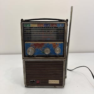 Może przedstawiać: Przenośne radio vintage marki Sonnet z brązową plastikową maskownicą głośnika, czarnym uchwytem do przenoszenia i długą srebrną anteną teleskopową. Panel sterowania zawiera grafikę mapy świata, trzy srebrne pokrętła i skale częstotliwości.