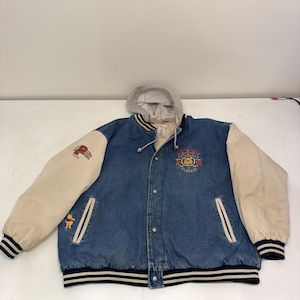 Op de afbeelding: Een blauwe spijkerjas in varsity-stijl met crèmekleurige mouwen en een grijze jersey capuchon. De voorkant heeft een geborduurde Winnie the Pooh Classic patch, terwijl de rechtermouw een letter P patch en een kleine Pooh-figuur bevat.