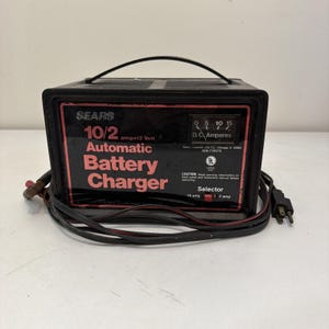 Puede incluir: Un cargador de batería automático negro Sears de 10/2 amperios y 12 voltios con asa de transporte. El panel frontal incluye un medidor analógico de amperios D.C., un selector rojo para 10 o 2 amperios y cables de alimentación y carga.