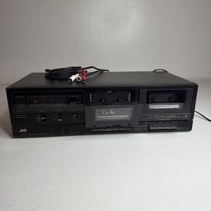 Puede incluir: Una pletina de casete doble estéreo JVC negra con dos compartimentos para casetes, uno de los cuales está abierto. El panel frontal incluye varios botones de control, un interruptor de encendido y un contador digital. Se adjunta un cable de alimentación negro con conectores RCA rojo y blanco.