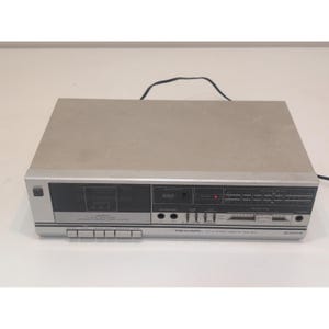 May include: A silver and gray Realistic SCT-23 stereo cassette deck. The deck has a cassette tape slot, various function buttons, and a display with a red power indicator. The front panel has the Realistic logo and model number.