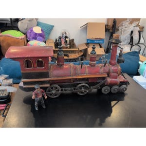 May include: A vintage model train in red and black. The train has a detailed design with multiple wheels, smokestacks, and a cabin. A small figurine of a train conductor stands in front of the train.