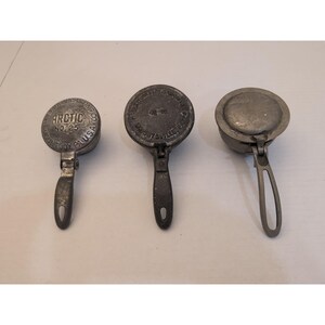 May include: Three vintage metal ice cream scoops or moulds with hinged handles. The leftmost tool is marked Arctic No. 25 Mount Joy, PA, U.S.A. The middle tool features circular text and a central hinge. The rightmost tool has a smooth, plain metal surface.