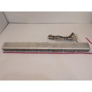 May include: A silver rectangular light bar with multiple small lights. A pink measuring tape is placed along the bottom, showing measurements in inches. A white power cord is coiled on top.