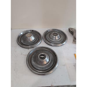 Könnte beinhalten: Drei Vintage-Radkappen aus Chrom mit radialem Design und zentralem Emblem. Die Radkappen sind kreisförmig, mit einer Reihe konzentrischer Rippen, die von der Mitte ausgehen. Das Emblem in der Mitte ist ein schwarz-silbernes Chevrolet-Logo.