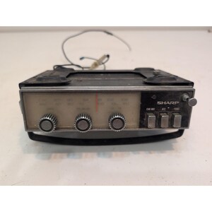 Puede incluir: Una radio de coche Sharp vintage con carcasa metálica negra rectangular, pantalla de frecuencia analógica central con aguja roja, tres mandos de control plateados circulares y tres botones rectangulares etiquetados CAR ANT, AFC y TONE.
