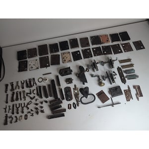 Op de afbeelding: Een grote verzameling diverse vintage hardware-onderdelen, waaronder rechthoekige metalen slotplaten, diverse deurknoppen, versierde sleutelplaatjes, scharnieren, grendels en kleine ijzeren bevestigingsmiddelen, verspreid over een oppervlak.