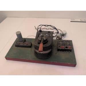 May include: Vintage American Flyer train control panel. The panel is black with a red and green base. It features a control lever, buttons, and a small grey cylinder. The words "American Flyer" are visible on the panel.