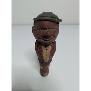 May include: A carved wooden bottle stopper featuring a figure with a brown face, a green hat, and a red jacket with a white vest. The figure has a carved mouth line and a cork base. The wood shows signs of wear and paint chipping on the surface.